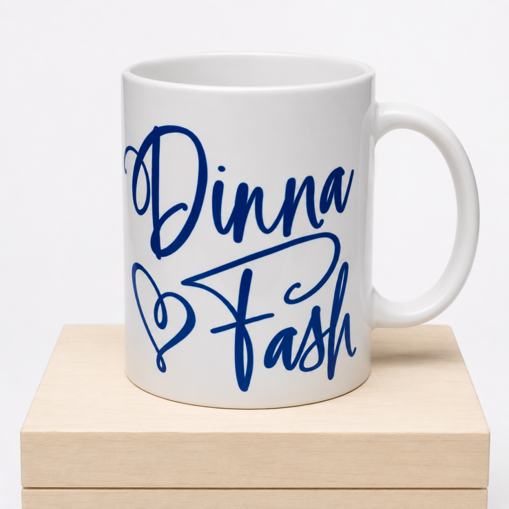 White ceramic mug with blue “Dinna Fash” script design displayed on a light wooden pedestal.